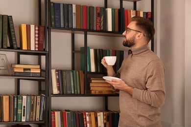 Photo of Young man with cup of coffee near shelves in home library