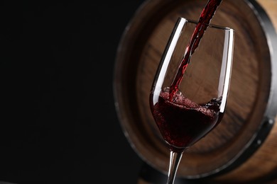 Photo of Pouring red wine into glass near wooden barrel against black background, closeup. Space for text