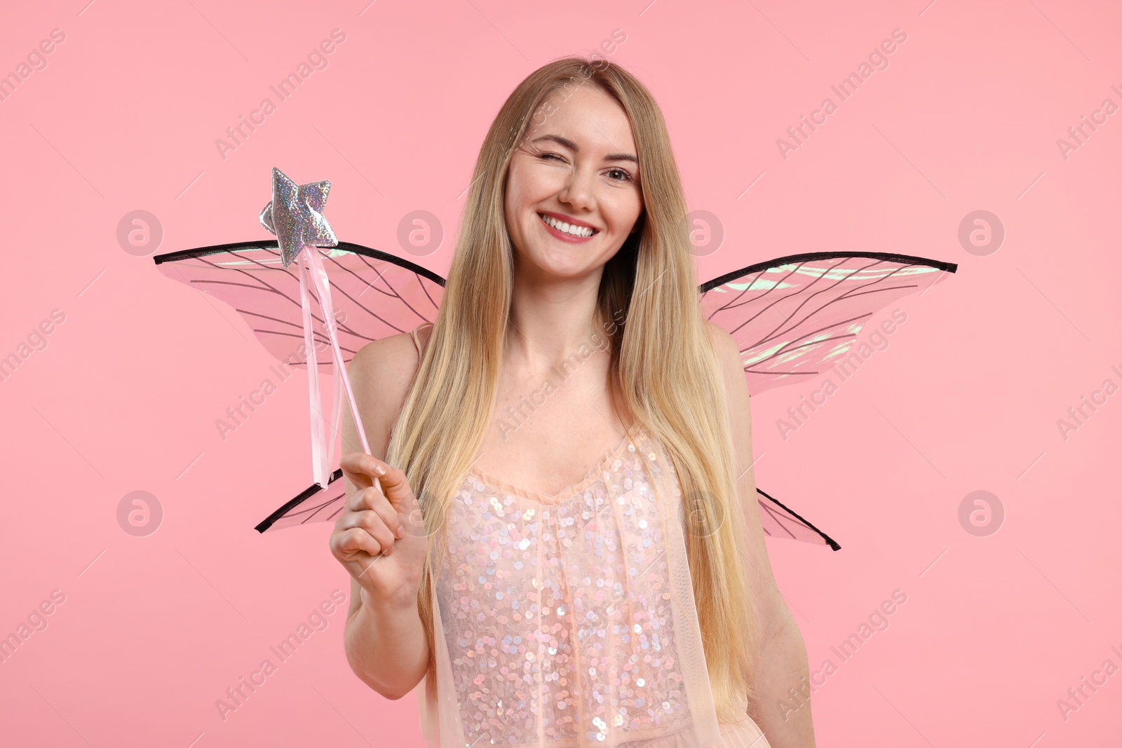 Photo of Beautiful girl in fairy costume with wings and magic wand winking on pink background