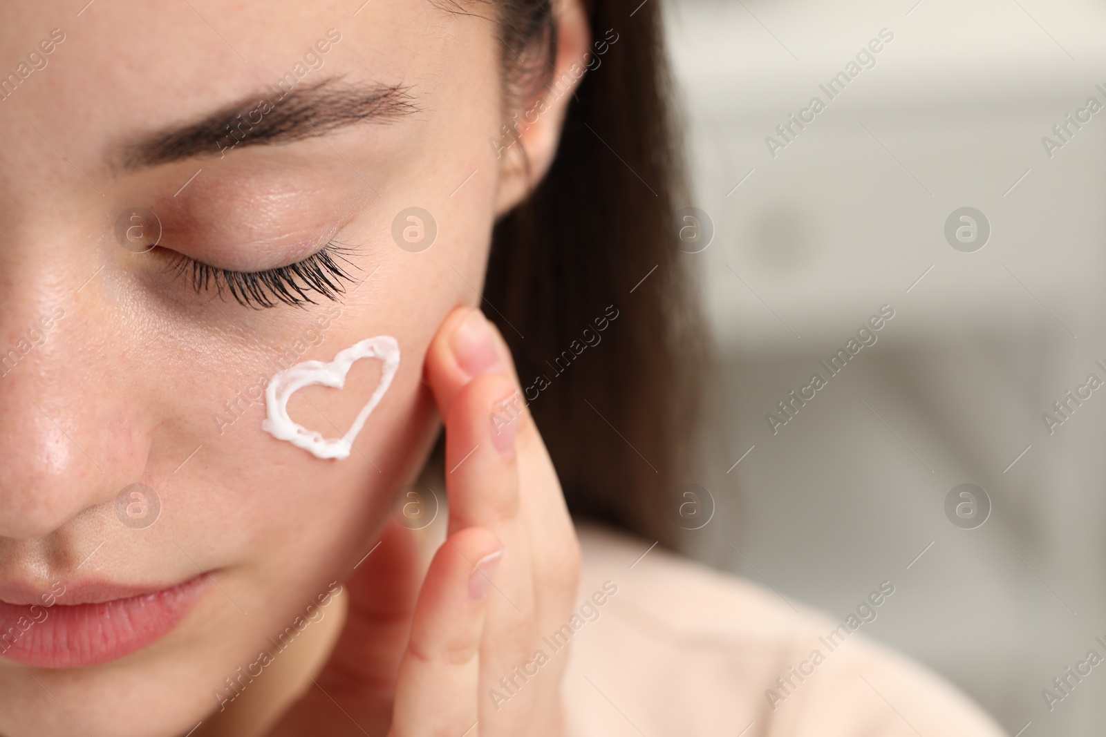 Photo of Young woman with dry skin and heart made of cream on her face indoors, closeup. Space for text