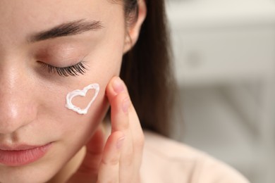 Young woman with dry skin and heart made of cream on her face indoors, closeup. Space for text