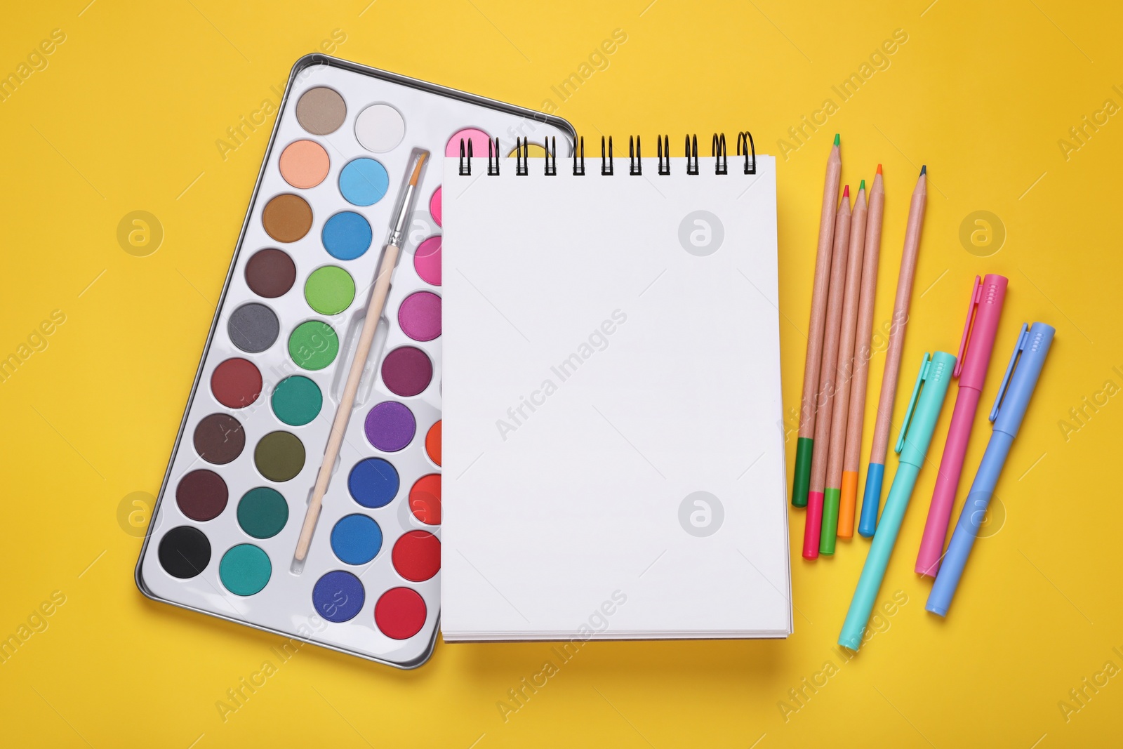 Photo of Watercolor palette, empty notebook, colorful pencils and markers on yellow background, flat lay. Space for text