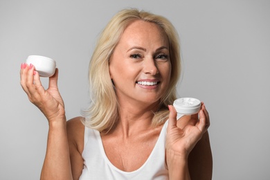 Portrait of beautiful mature woman with perfect skin holding jars of cream on grey background