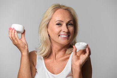 Photo of Portrait of beautiful mature woman with perfect skin holding jars of cream on grey background