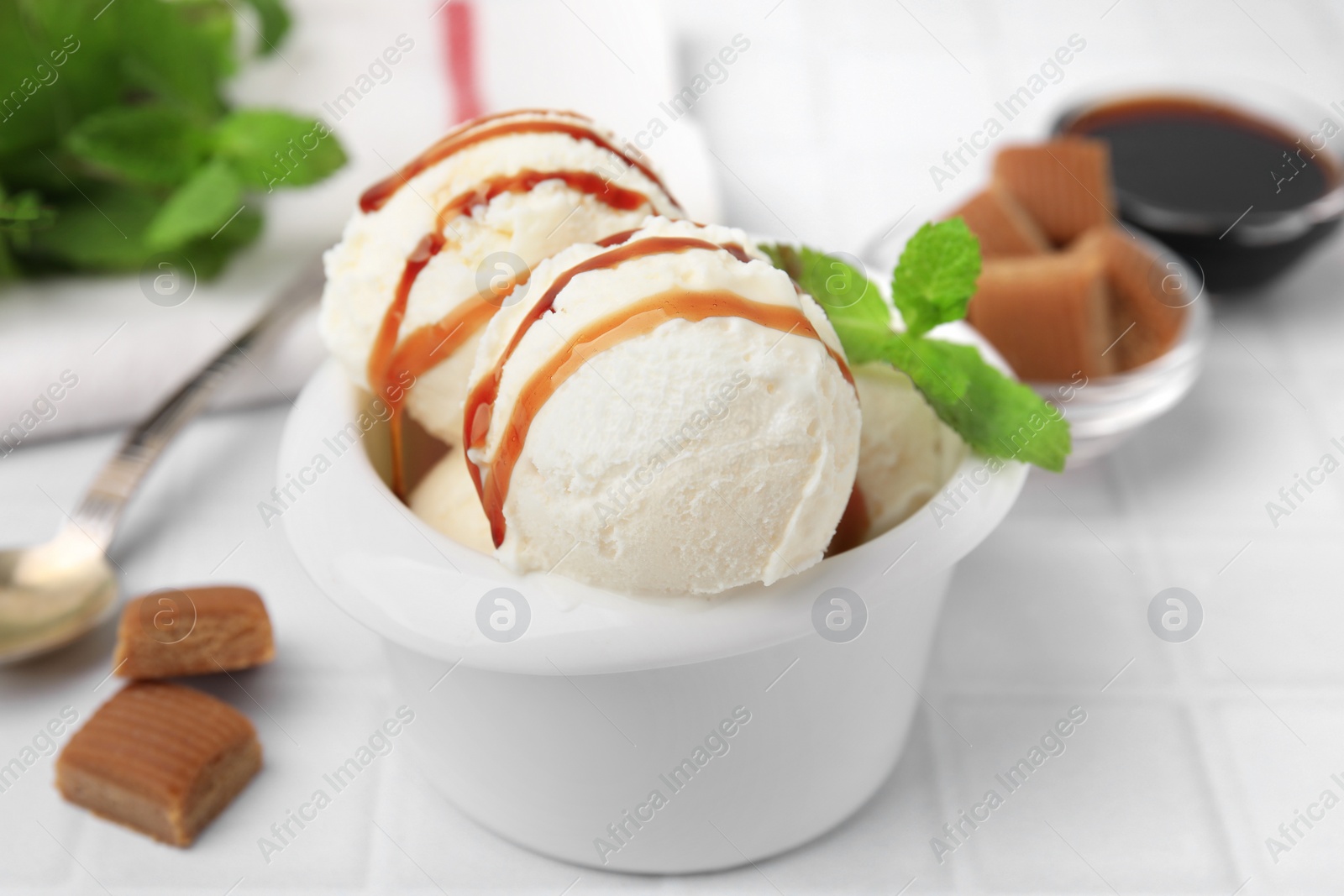 Photo of Scoops of tasty ice cream with mint leaves, candies and caramel sauce on white tiled table, closeup