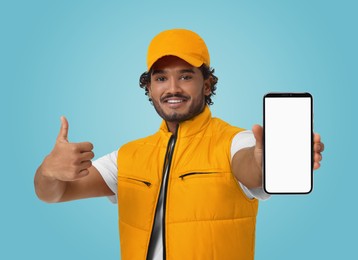 Image of Happy courier holding smartphone with empty screen and showing thumbs up on light blue background