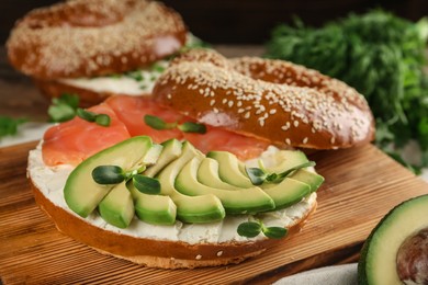 Photo of Delicious bagel with cream cheese, salmon, avocado and microgreens on wooden board, closeup