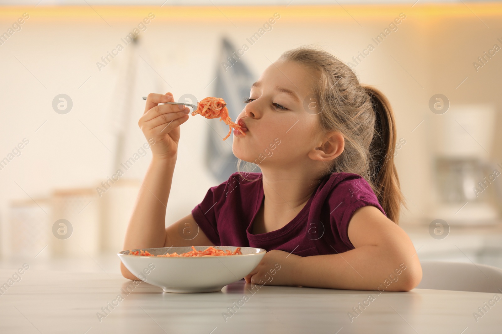 Photo of Cute little girl eating tasty pasta at table in kitchen