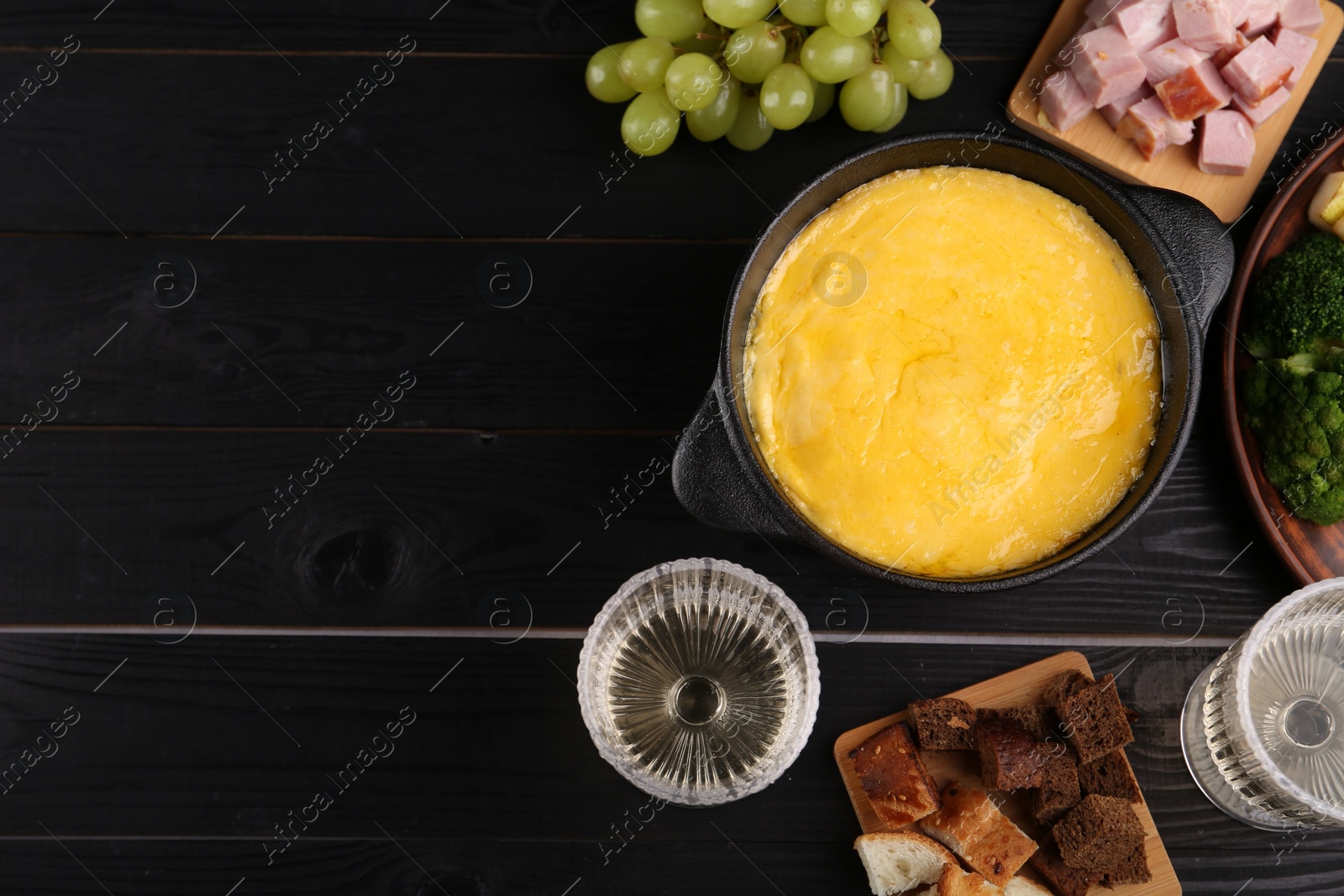 Photo of Fondue pot with melted cheese and different products on black wooden table, flat lay. Space for text