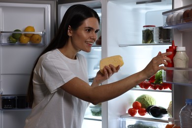 Young woman taking cheese out of refrigerator at night