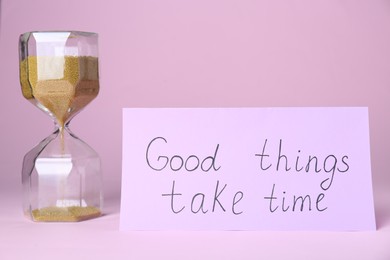 Photo of Card with phrase Good Things Take Time and sand clock on pink background. Motivational quote