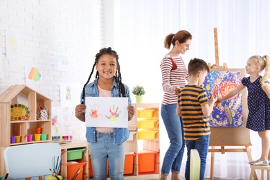 Cute little child showing sheet of paper with colorful hand prints indoors. Painting lesson