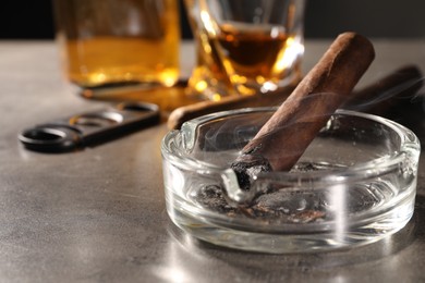 Photo of Smoldering cigar, ashtray and whiskey on grey table, closeup. Space for text