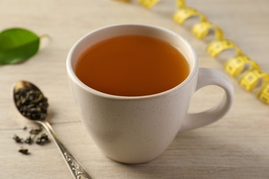 Cup of herbal diet tea and measuring tape on light wooden table, closeup. Weight loss concept