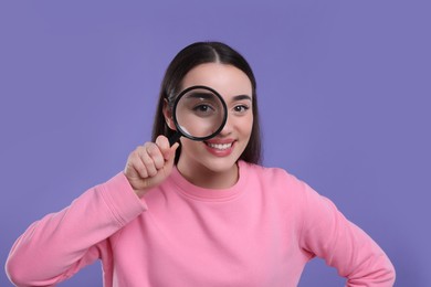 Happy young woman looking through magnifier glass on purple background
