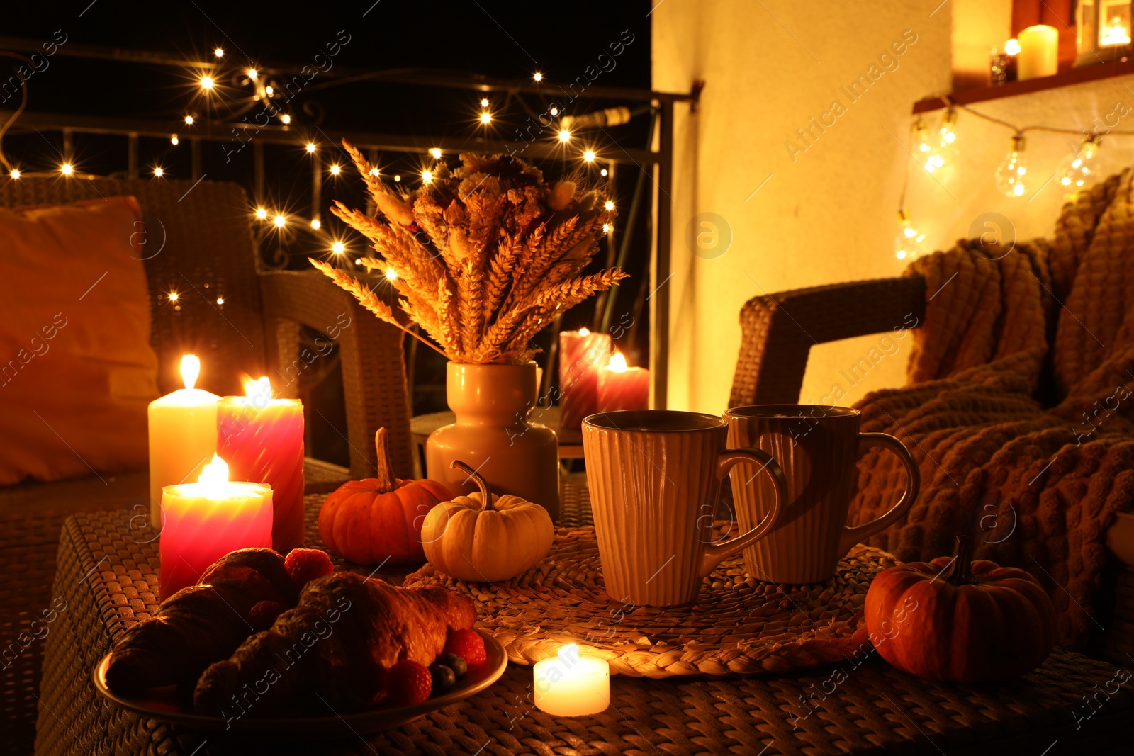 Photo of Rattan furniture, cups, fairy lights, burning candles and other autumn decor on outdoor terrace at night