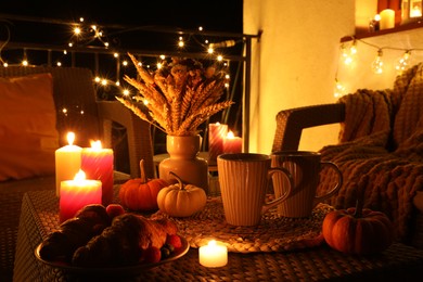 Photo of Rattan furniture, cups, fairy lights, burning candles and other autumn decor on outdoor terrace at night