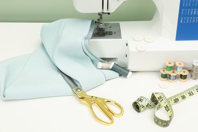 Photo of Sewing machine with fabric, scissors, measuring tape and threads on white table, closeup