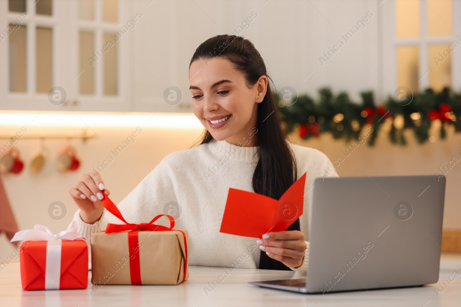 Photo of Celebrating Christmas online with exchanged by mail presents. Smiling woman holding greeting card and opening gift during video call at home