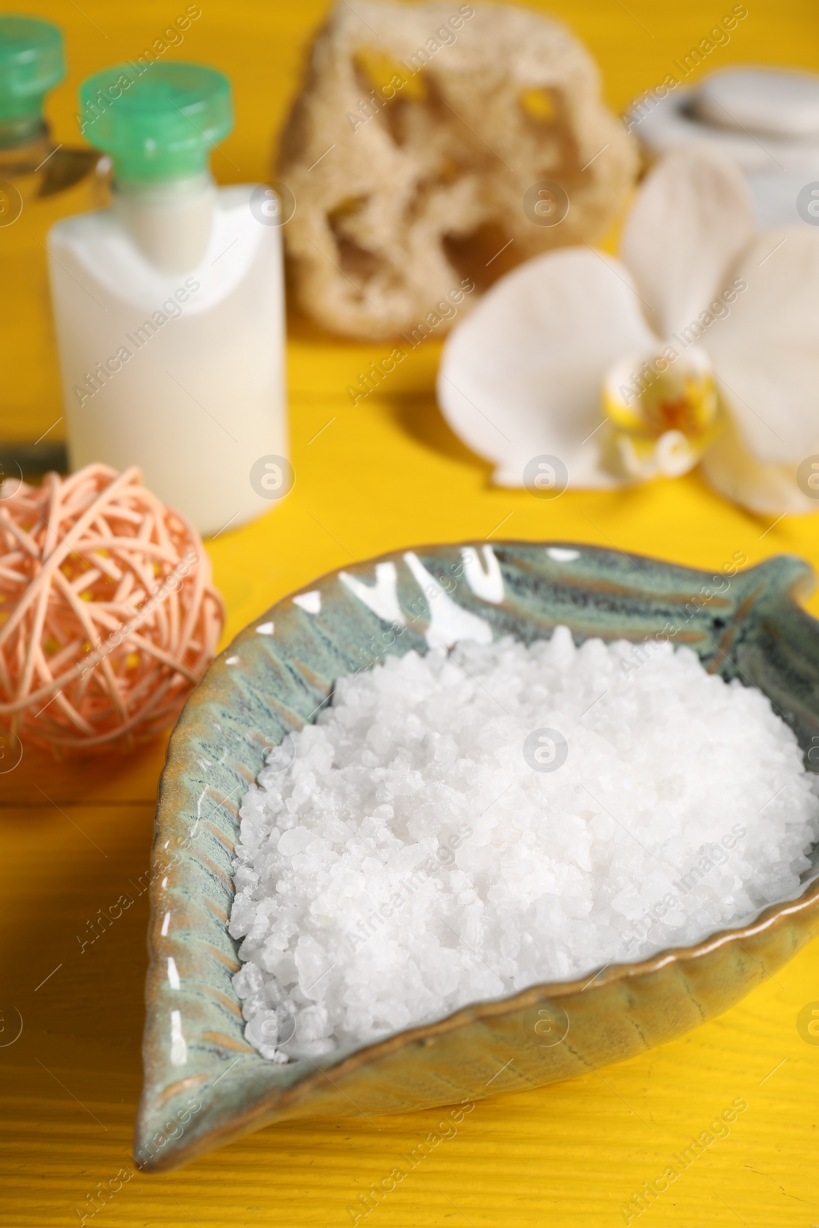 Photo of Natural sea salt in bowl and other spa products on yellow wooden table, closeup