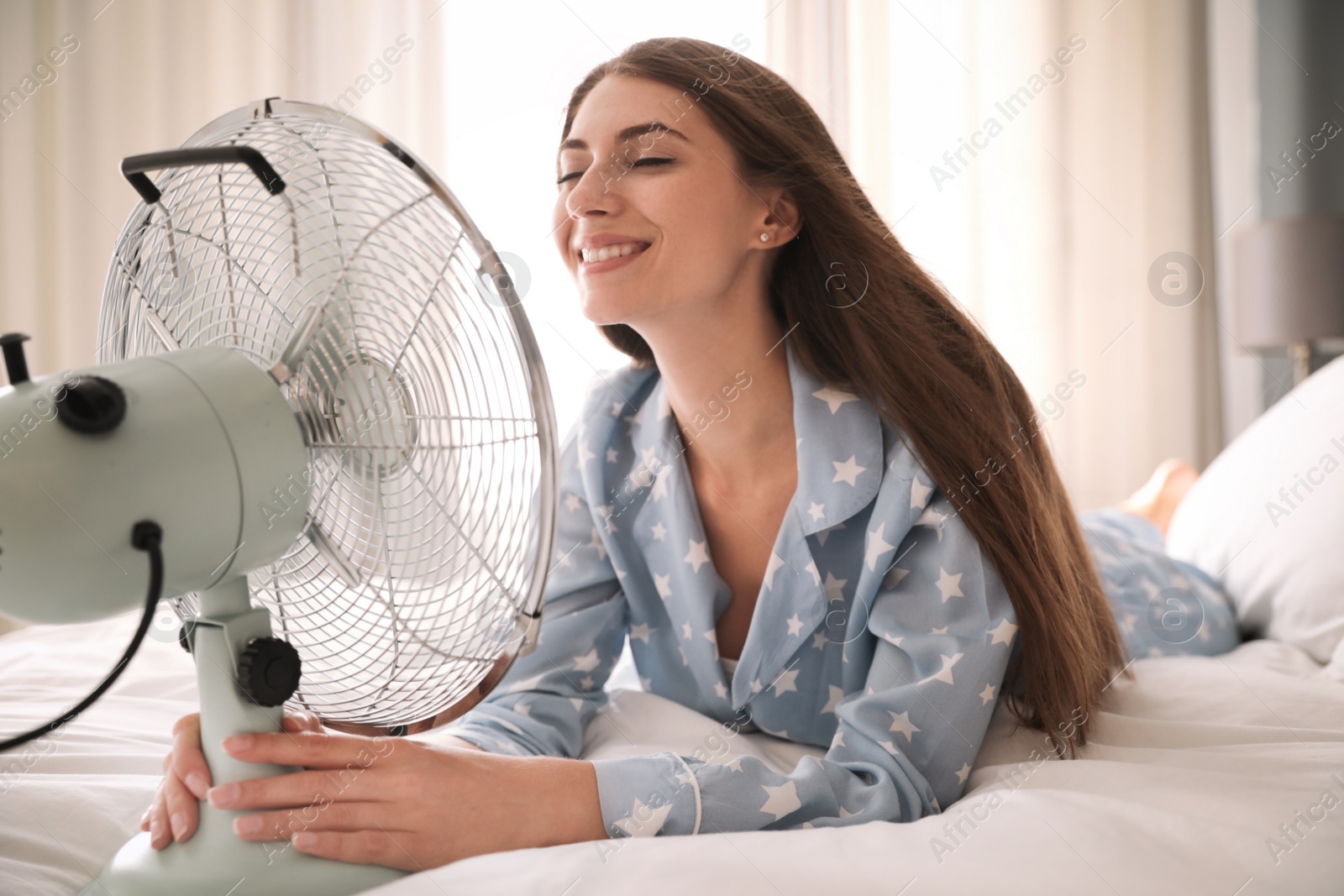 Photo of Woman enjoying air flow from fan on bed in room. Summer heat