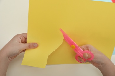 Photo of Child cutting color paper with plastic scissors at table, closeup