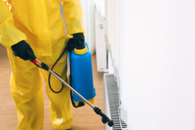 Pest control worker in protective suit spraying pesticide indoors, closeup. Space for text