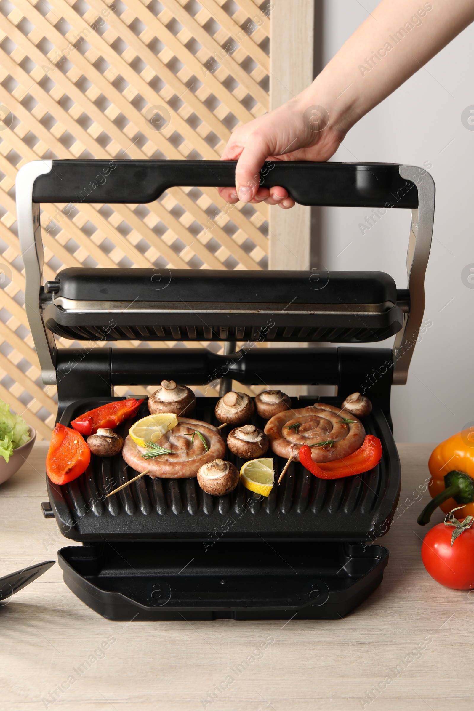 Photo of Woman cooking homemade sausages with bell peppers and mushrooms on electric grill at wooden table, closeup