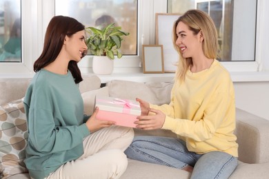 Photo of Smiling young woman presenting gift to her friend on sofa at home