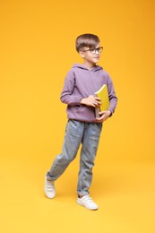 Photo of Cute schoolboy in glasses holding books and walking on orange background
