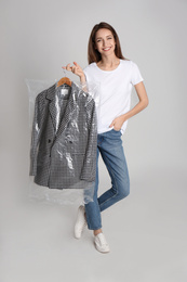 Photo of Young woman holding hanger with jacket in plastic bag on grey background. Dry-cleaning service