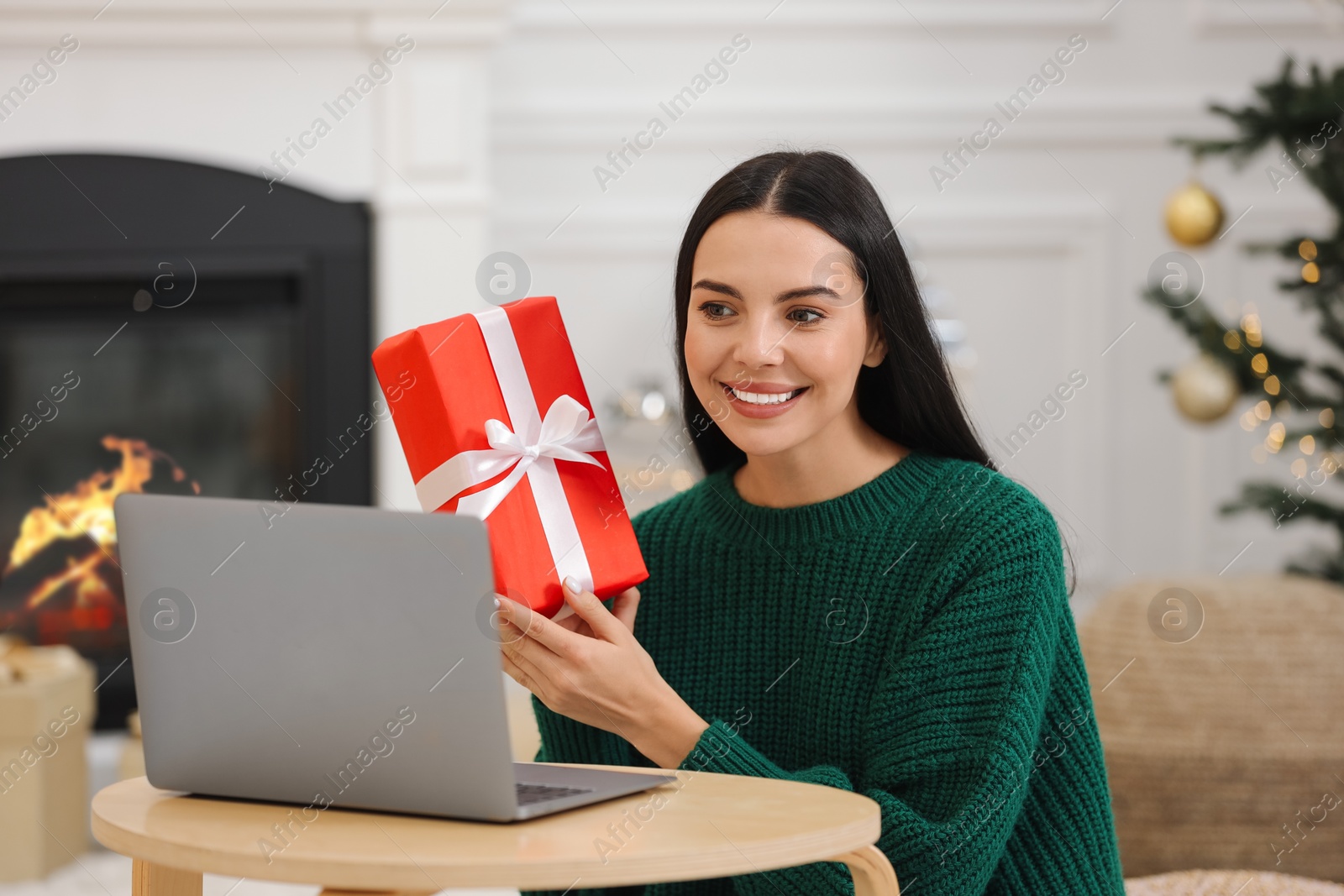 Photo of Celebrating Christmas online with exchanged by mail presents. Smiling woman with gift box during video call on laptop at home