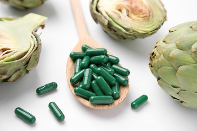 Photo of Spoon with pills and fresh artichokes on white background, closeup