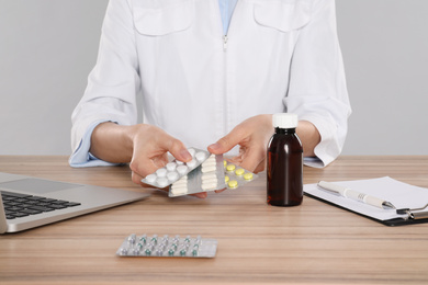 Professional pharmacist with pills and laptop at table against light grey background, closeup