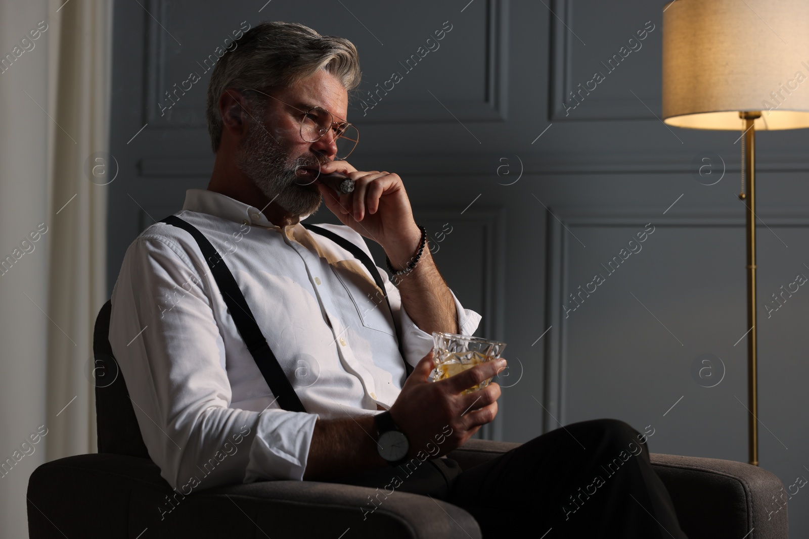 Photo of Bearded man with glass of whiskey smoking cigar in armchair indoors