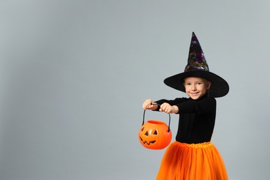 Photo of Cute little girl with pumpkin candy bucket wearing Halloween costume on grey background, space for text