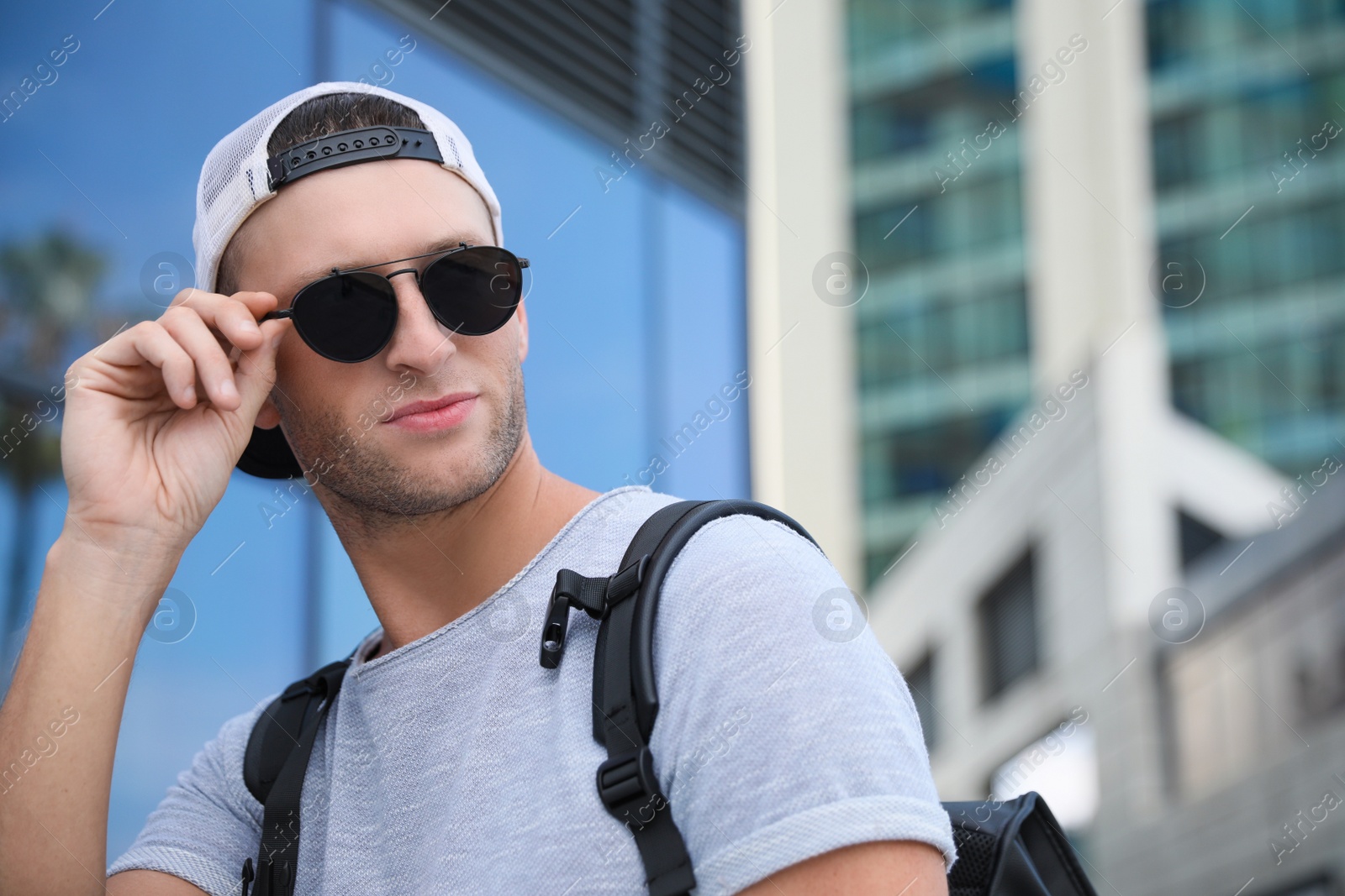 Photo of Handsome young man with stylish sunglasses and backpack near building outdoors, space for text