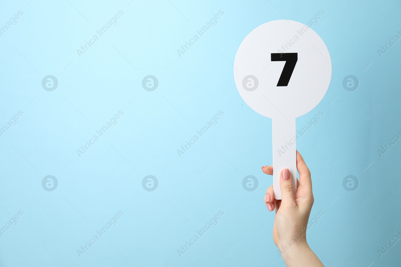 Photo of Woman holding auction paddle with number 7 on light blue background, closeup. Space for text