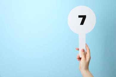 Photo of Woman holding auction paddle with number 7 on light blue background, closeup. Space for text