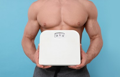 Athletic man holding scales on light blue background, closeup. Weight loss concept