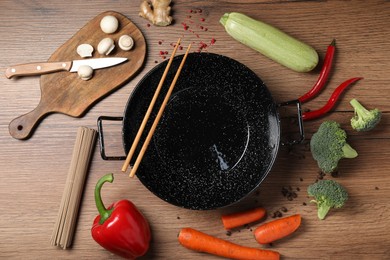 Photo of Empty iron wok surrounded by raw ingredients on wooden table, flat lay