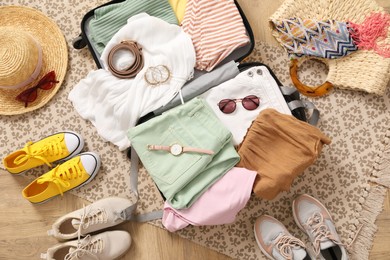 Photo of Open suitcase full of clothes and summer accessories near shoes on floor, top view