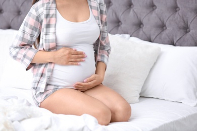 Young pregnant woman sitting on bed and touching her belly at home, closeup