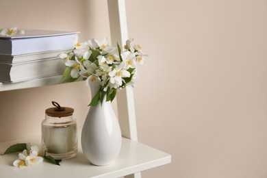Photo of Bouquet of beautiful jasmine flowers in vase and candle on shelving unit near beige wall indoors, space for text