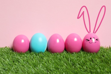 Image of One egg with drawn face and ears as Easter bunny among others on green grass against pink background