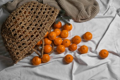 Photo of Stylish wicker bag with ripe tangerines on white bedsheet, flat lay