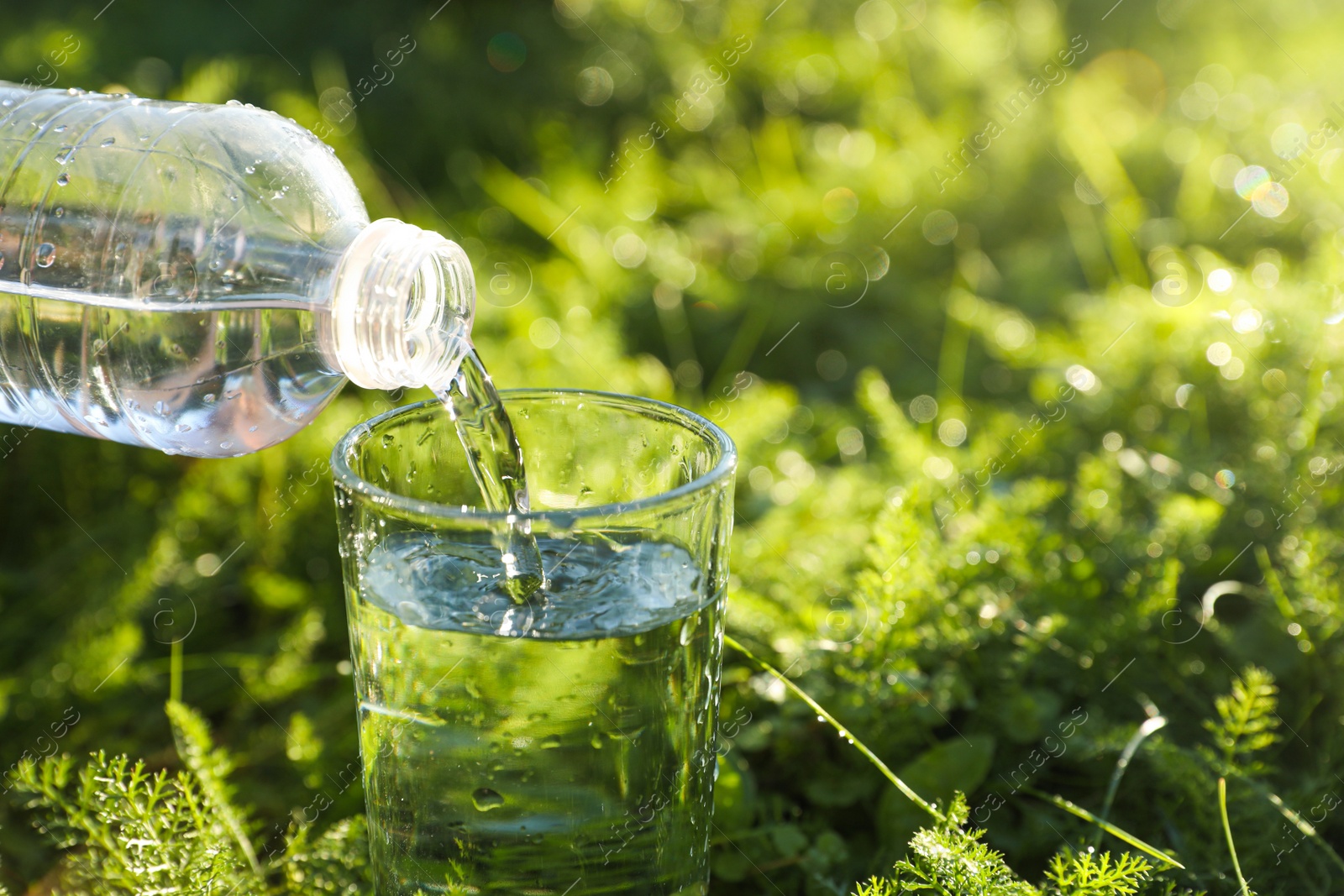 Photo of Pouring fresh water from bottle into glass on green grass outdoors, closeup. Space for text