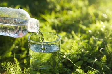 Pouring fresh water from bottle into glass on green grass outdoors, closeup. Space for text