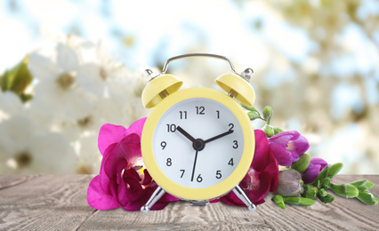 Image of Yellow alarm clock and flowers on table against blurred background. Spring time