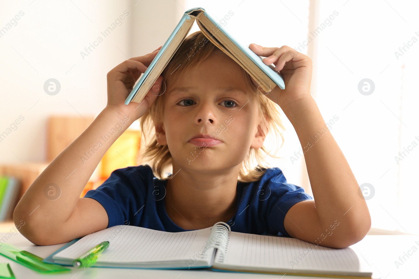 Photo of Bored little boy with book on his head doing homework at table indoors